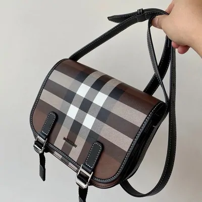 A check pattern flap crossbody bag with a leather strap, held up against a plain wall