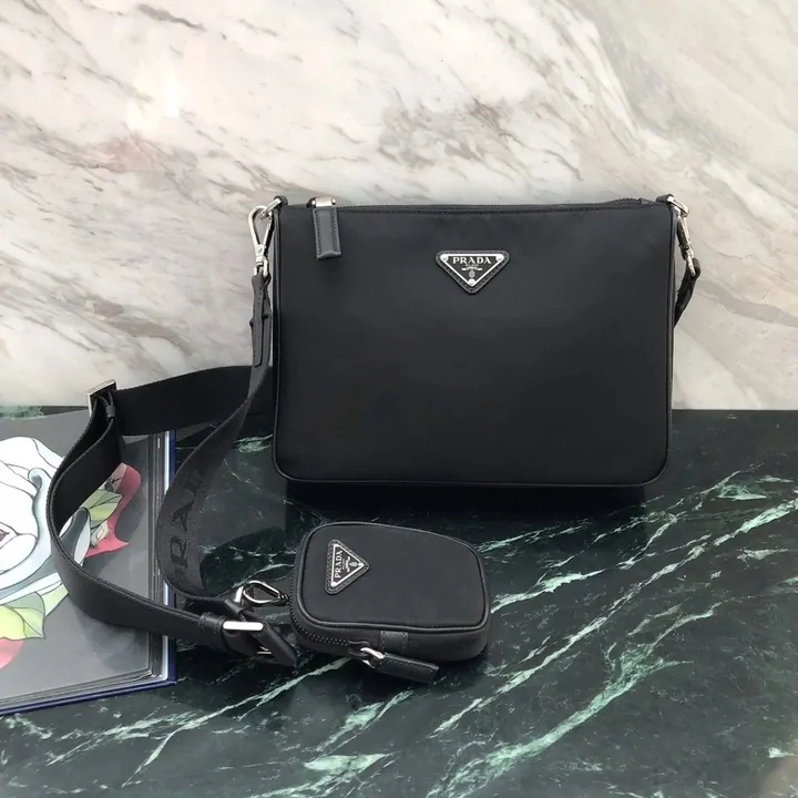 A black nylon crossbody bag with a small matching pouch clipped to the strap, photographed on a marble surface