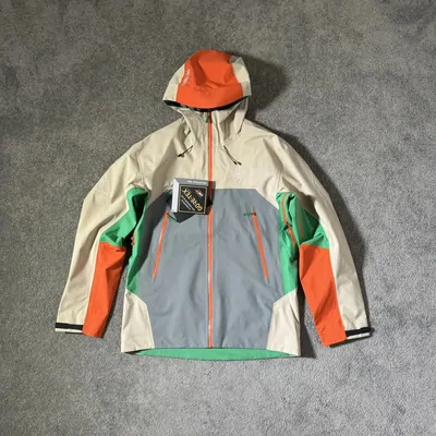 A colour blocked hooded shell jacket laid flat on grey concrete with its hang tag still attached