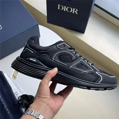 A black low top sneaker with a thick white sole, held up in one hand in front of its box