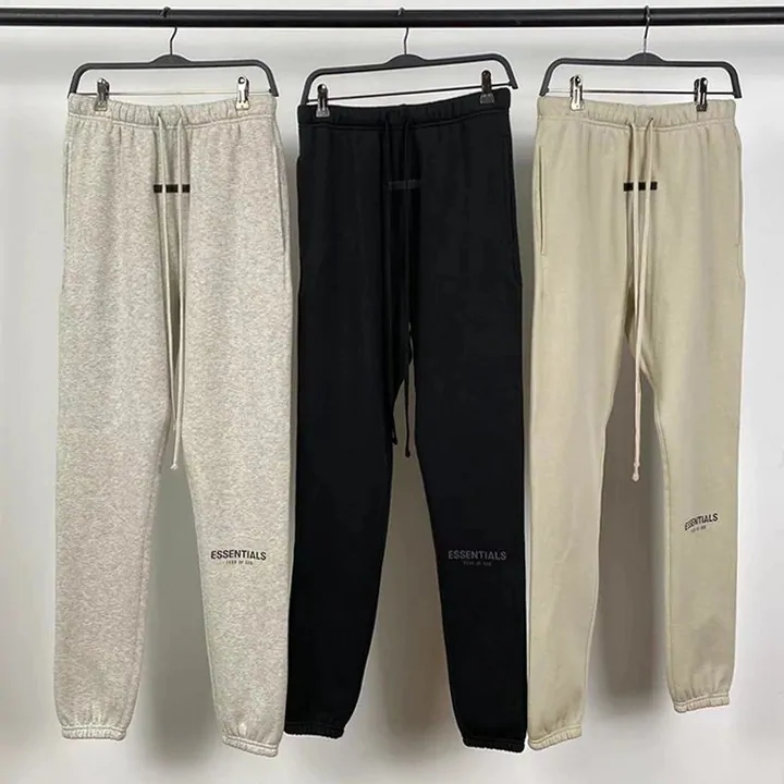Three pairs of jogging bottoms in grey, black and sand hanging side by side on a rail, each with the same chest logo at the hip