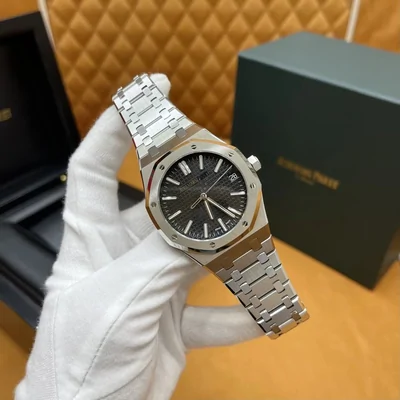 A steel bracelet watch with an octagonal bezel, held in a white gloved hand beside its case