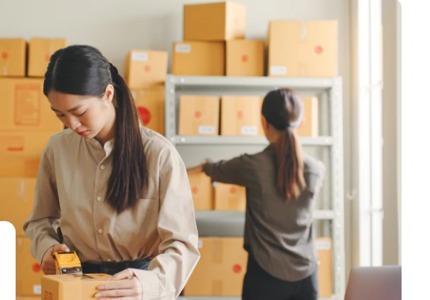 A warehouse worker taping a cardboard box shut at a wooden bench, with shelves of packed boxes behind her and a second worker in the background